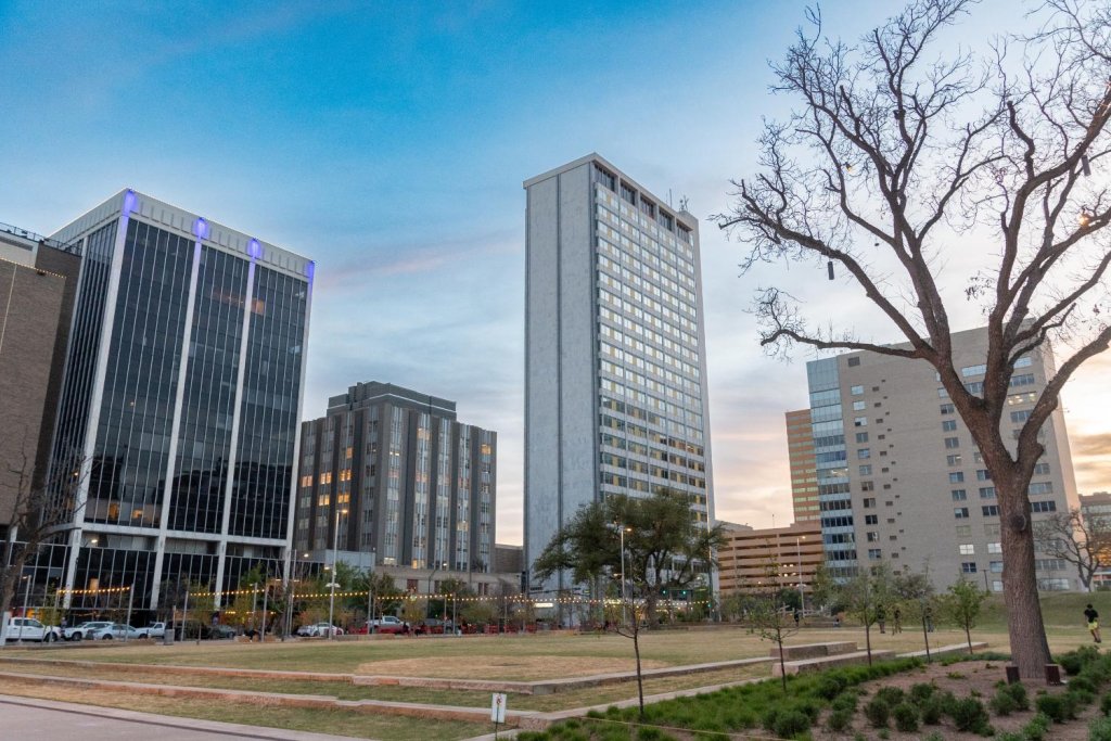 View of Centennial Park in downtown Midland, Texas, with modern high-rise buildings, open green space, and string lights at sunset.