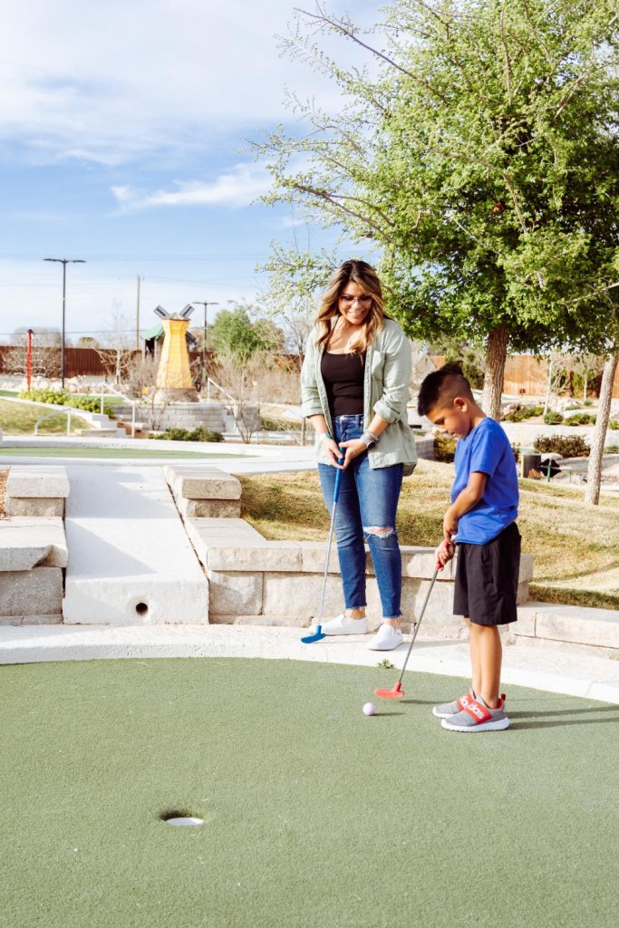 Woman and young boy playing a round of mini golf on a sunny day at Green Acres Mini Golf in Midland, Texas, surrounded by trees and playful course features.