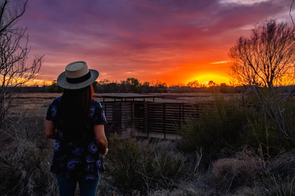 Woman in a straw hat watching a vibrant sunset over the wetlands at I-20 Wildlife Preserve in Midland, Texas, surrounded by native brush and silhouetted trees.