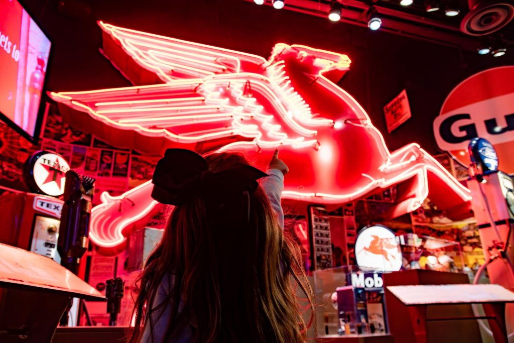 Young girl pointing at a large illuminated Pegasus neon sign inside the Permian Basin Petroleum Museum in Midland, Texas, surrounded by vintage gas station memorabilia.
